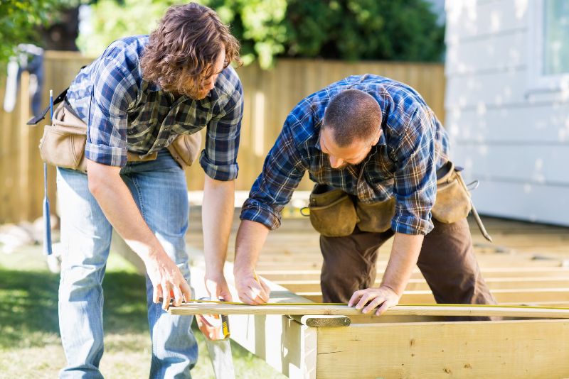 Two Men Laying Trying To Lay A Composite Deck While They Are Carrying Tool Belts On Their Waists